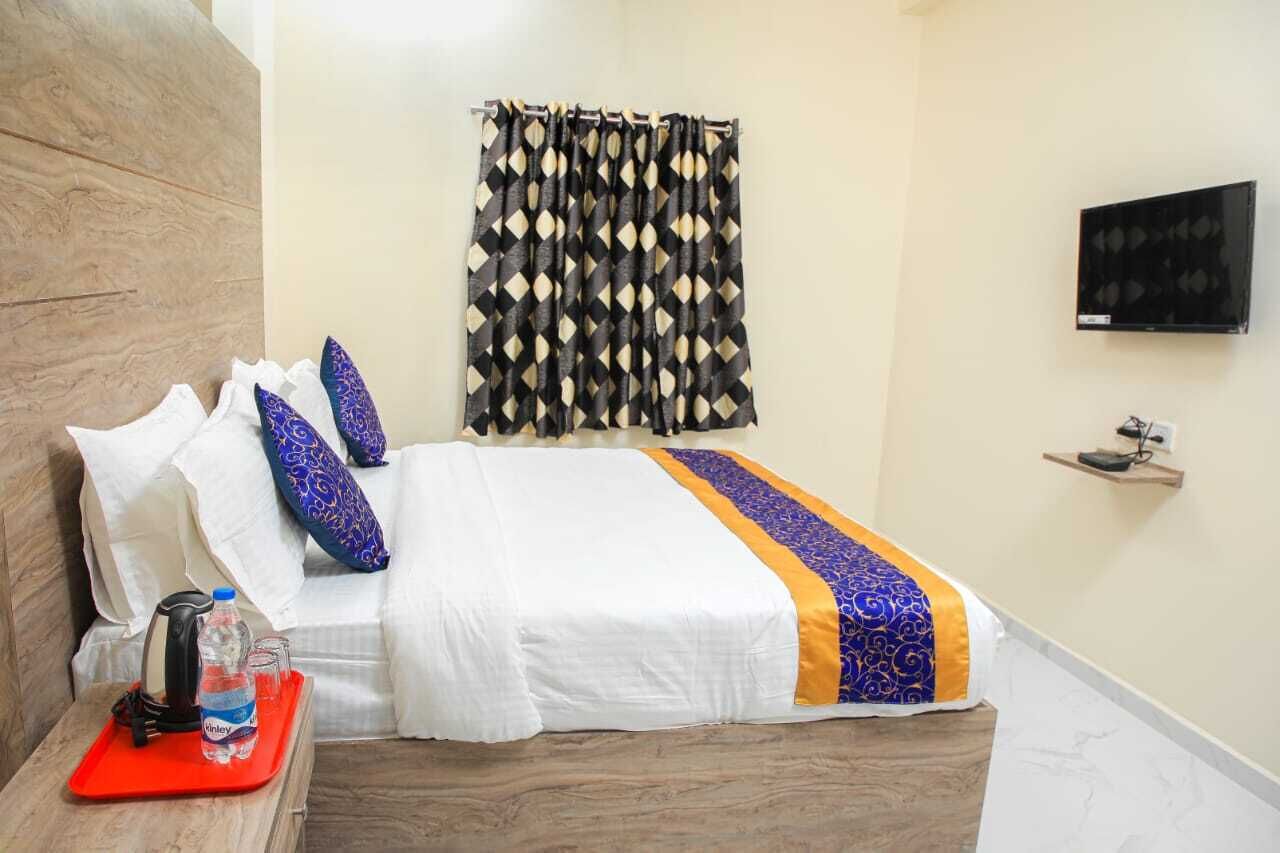 Habitación de hotel con cama, televisión, estante en la pared y ventana con cortinas estampadas.