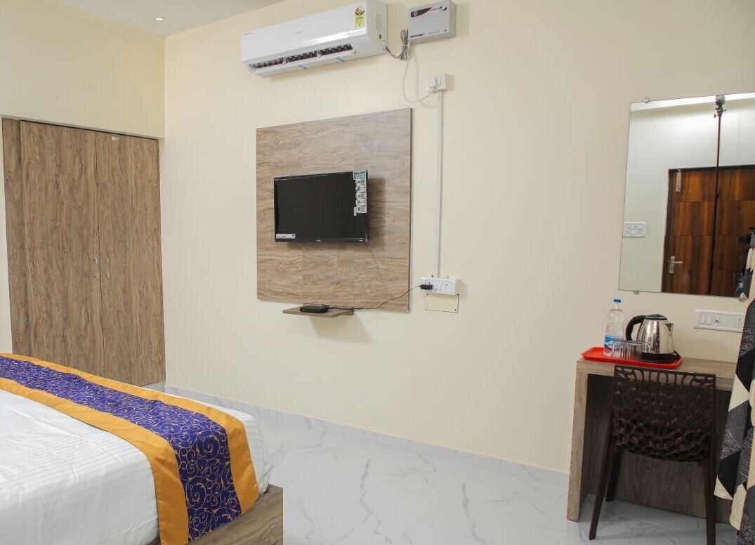 Habitación de hotel con cama, televisor montado en la pared, escritorio con hervidor y una silla.
