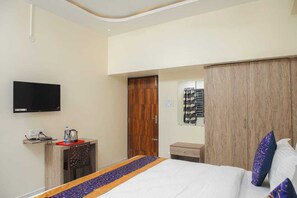 Ein Hotelzimmer mit Bett, Fernseher, Holzschrank, kleinem Tisch mit Wasserkocher und einer Wandablage.