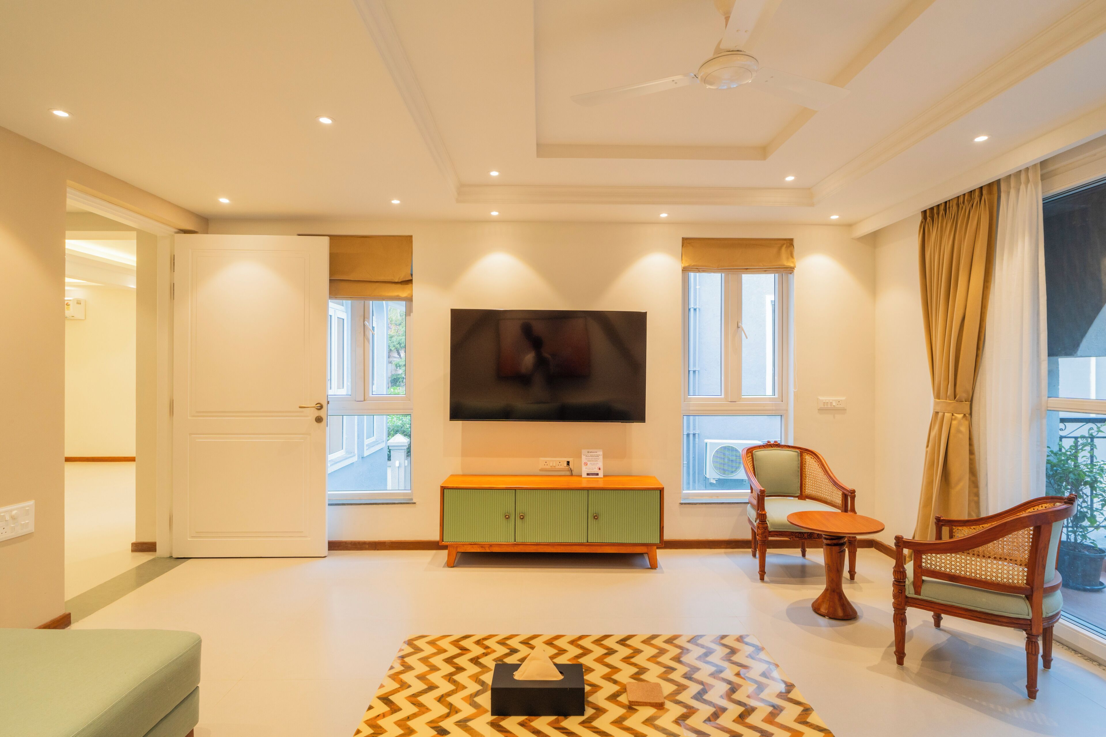 A modern living room with a flat-screen TV, a green console, two wooden chairs, and a patterned rug.