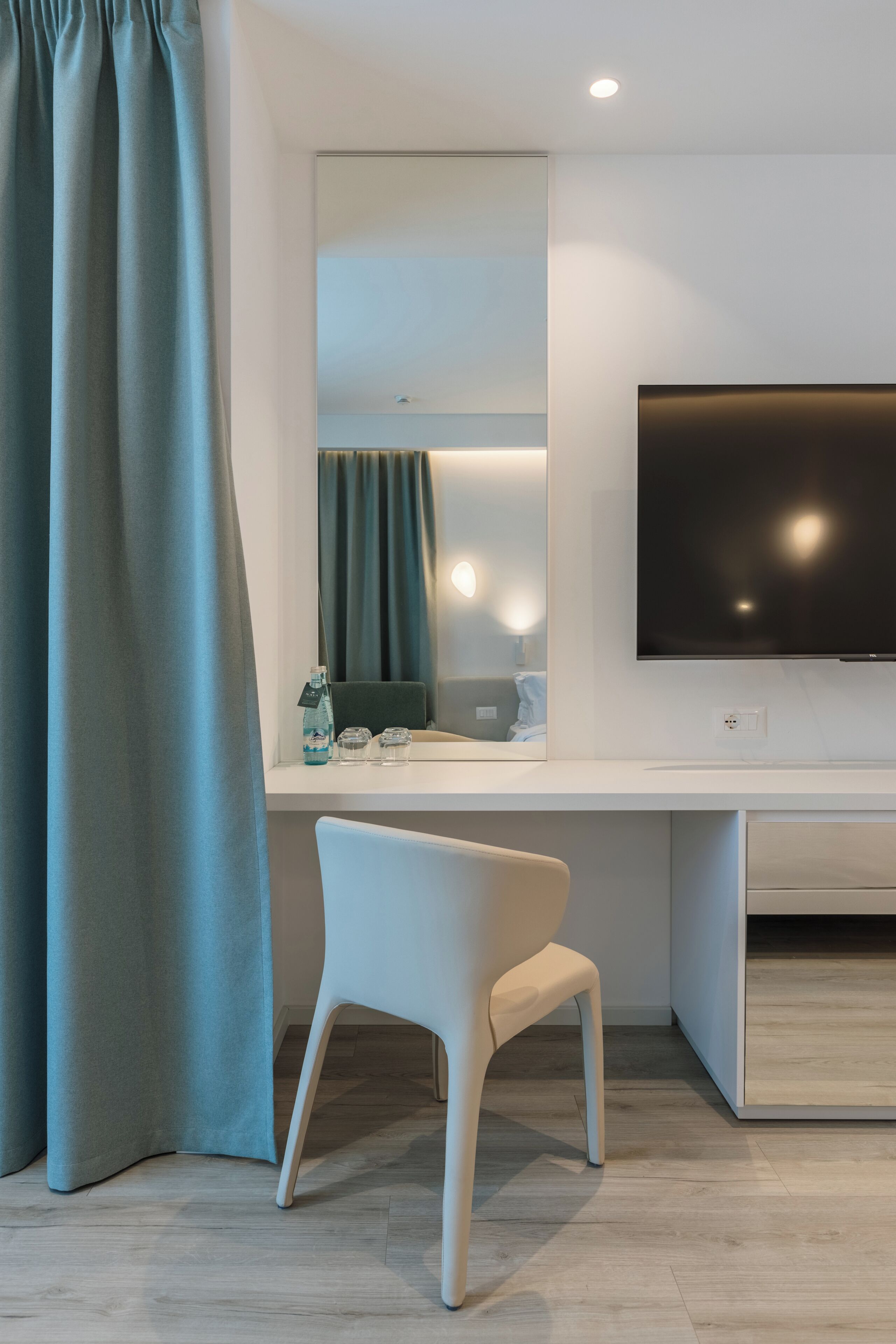 Ein modernes Hotelzimmer mit großem Flachbildfernseher, weißem Schreibtisch, Stuhl, Spiegel und Vorhang.