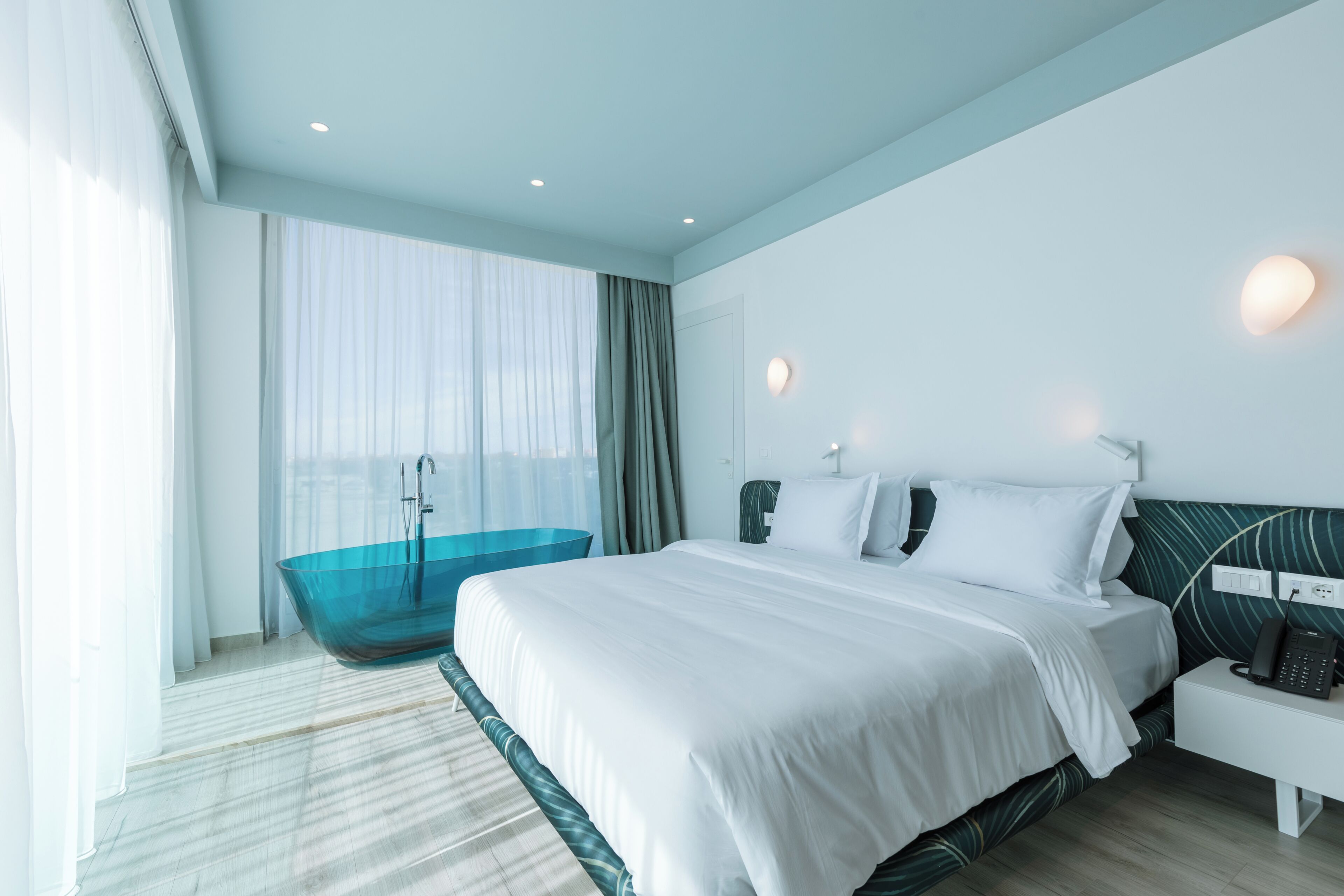 A modern hotel room with a large bed, a glass bathtub, and a bedside telephone.