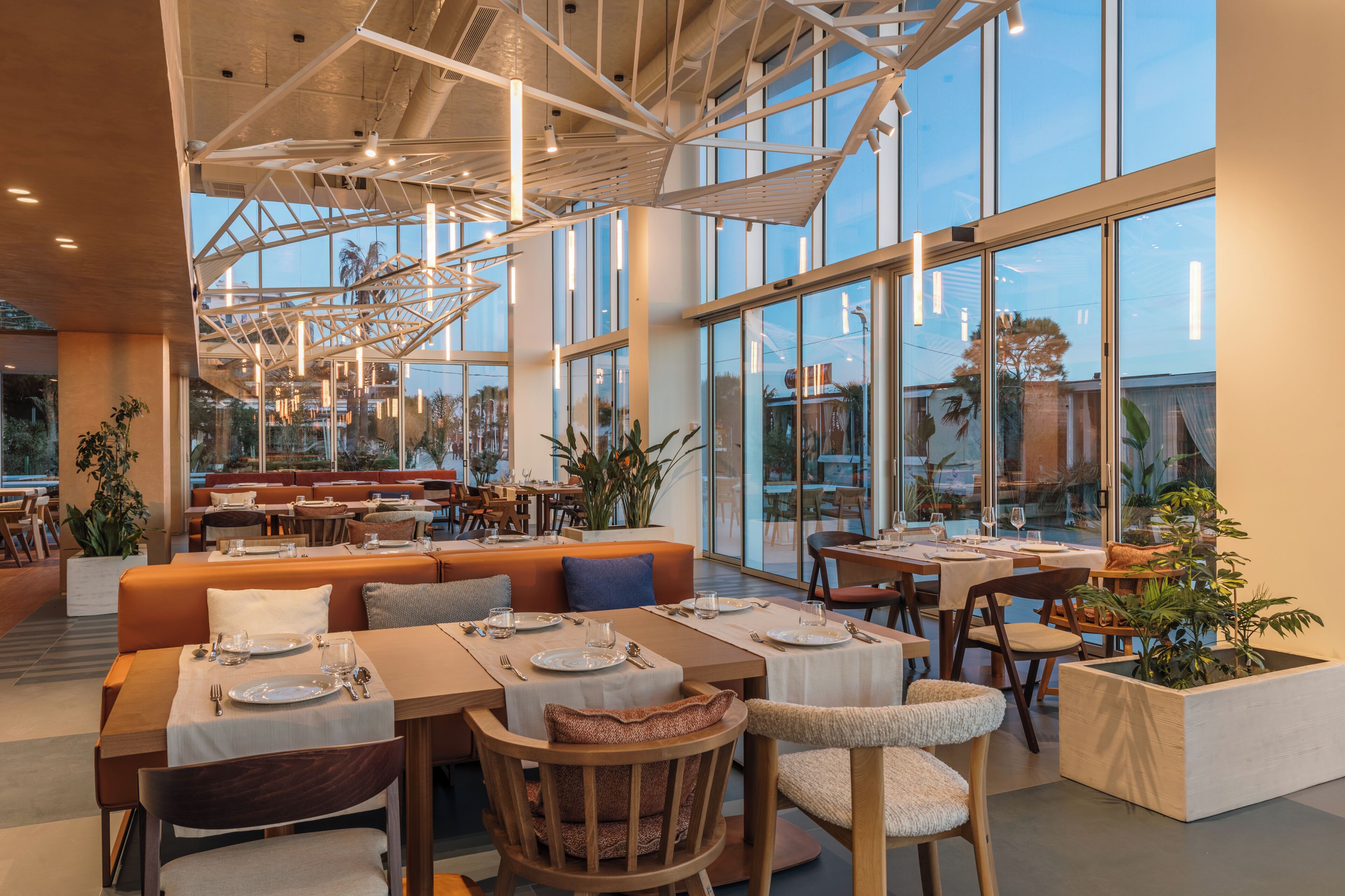 Een modern restaurant met hoge plafonds, grote ramen en diverse zitmogelijkheden, zoals tafels en hoekbanken.