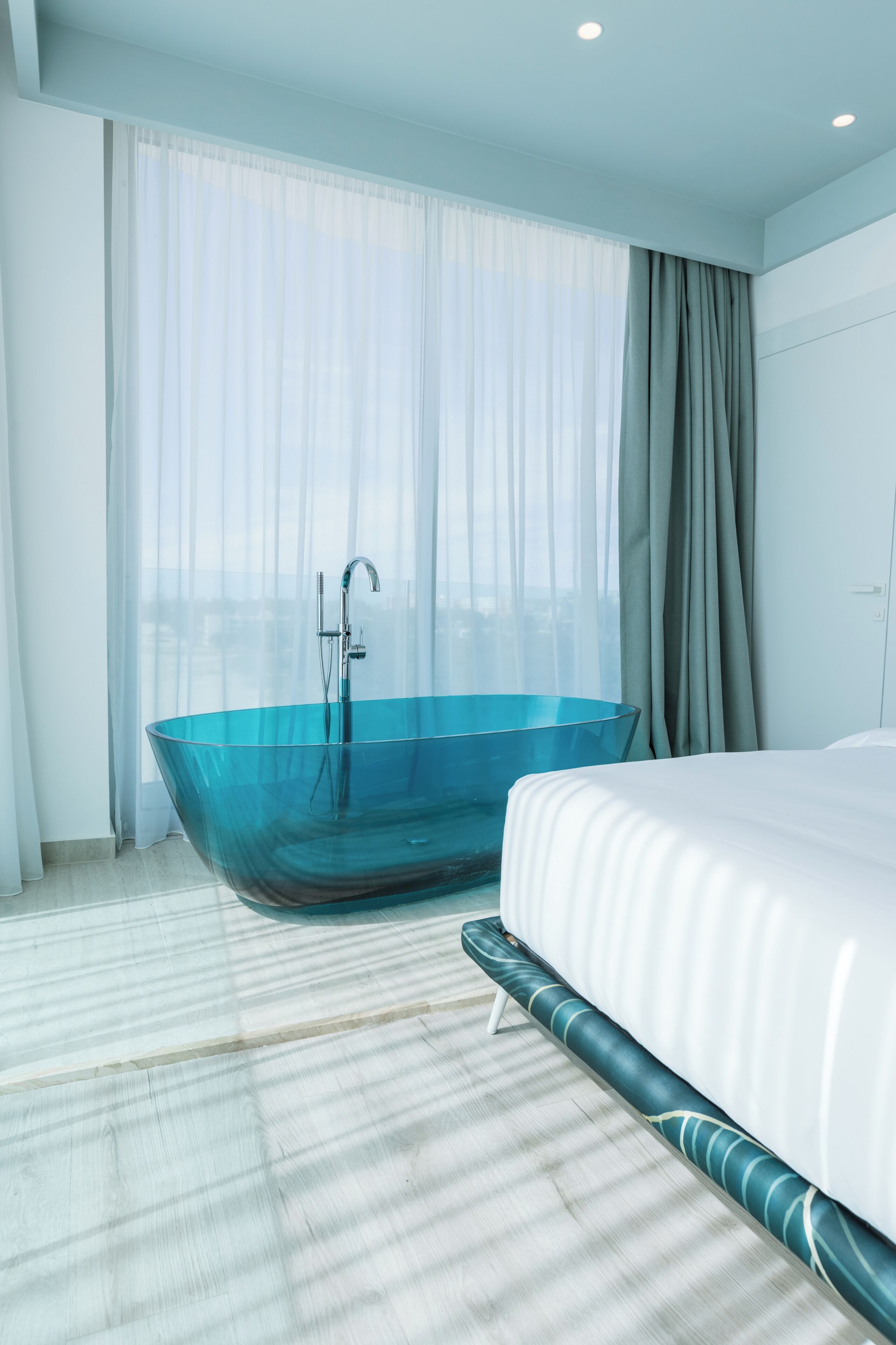 A modern hotel room with a large glass bathtub, a bed with white bedding, and a view through sheer curtains.