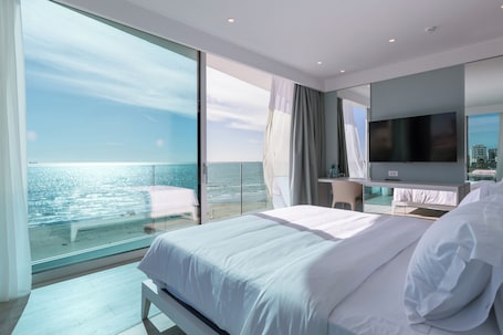 A modern hotel room with a large bed, a TV, and a balcony with a view of the sea.