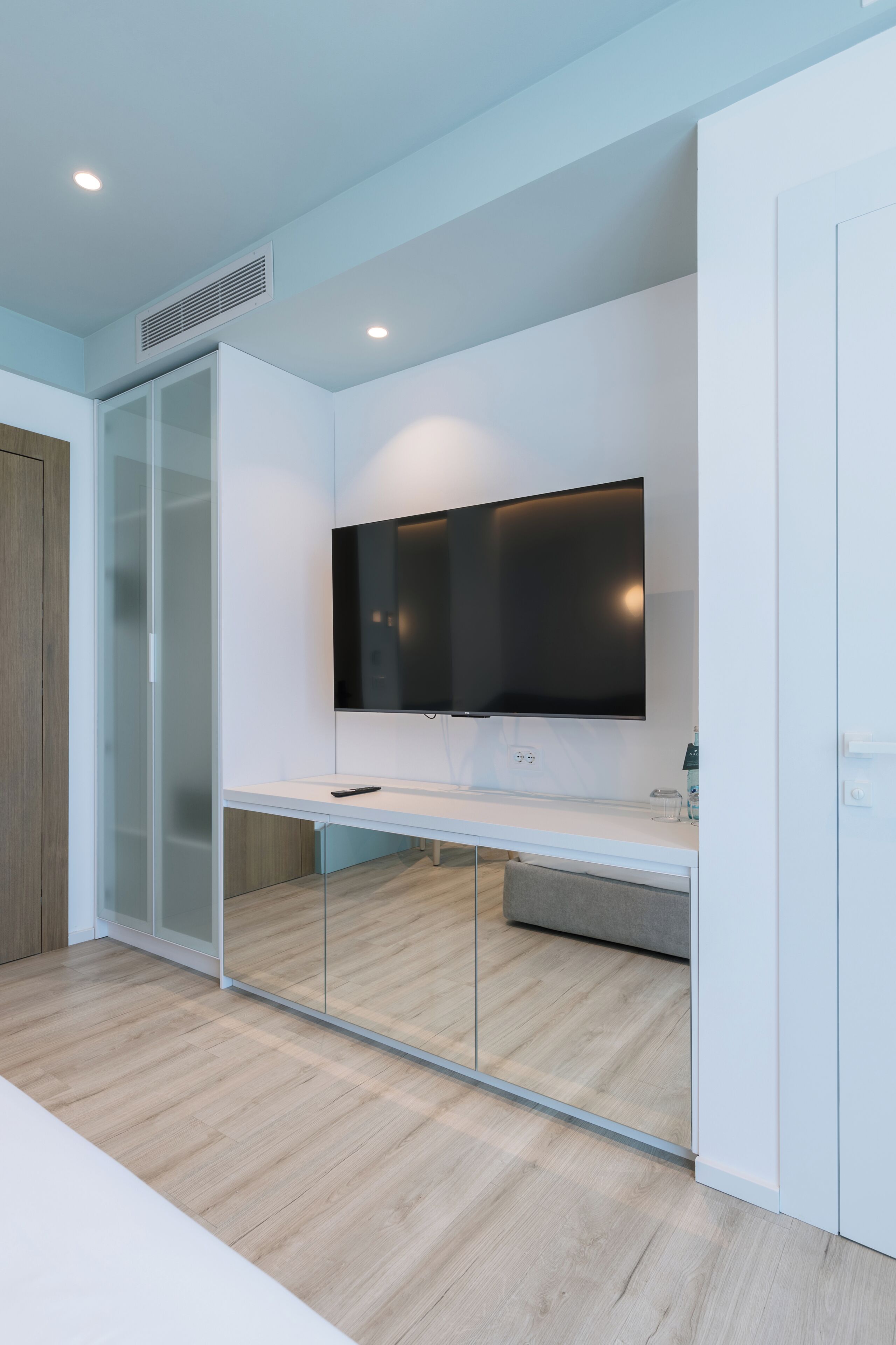 A modern room with a flat-screen TV mounted on the wall, a glass-fronted wardrobe, and a wooden floor.
