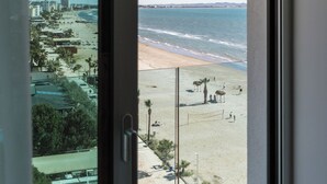 Vista desde una ventana a un lugar frente a la playa, con cielo despejado, arena dorada y un paisaje urbano al fondo.
