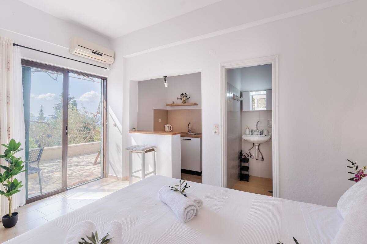 A bedroom with a bed, a small kitchenette, a bathroom, and a balcony with a chair.