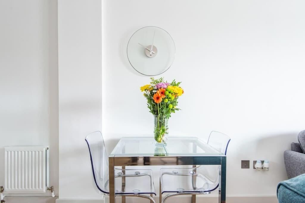 A modern dining area with a glass table, four chairs, a vase with flowers, and a wall clock.