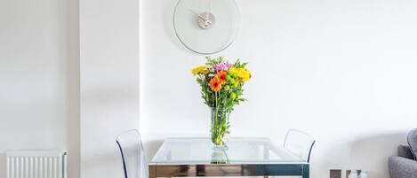 A modern dining area with a glass table, four chairs, a vase with flowers, and a wall clock.