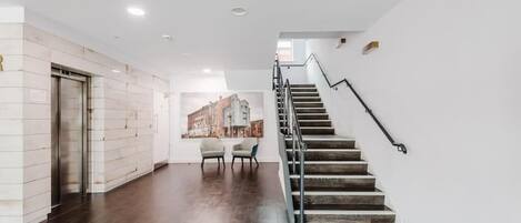 Un interior moderno con una escalera, un ascensor de pared y una zona de estar con sillas.