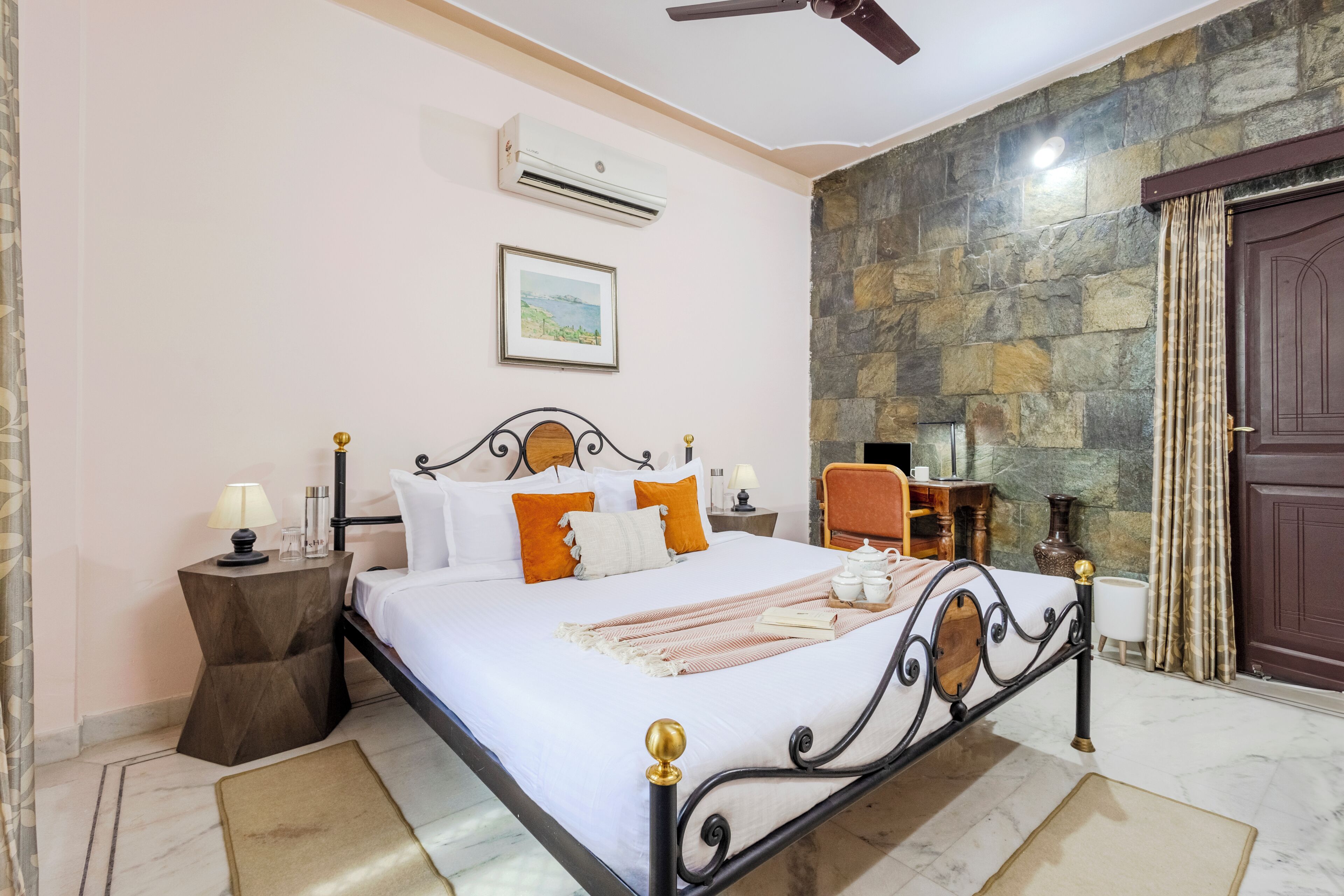 A neatly made bed with pillows and a blanket, a bedside table with a lamp, a framed picture on the wall, a stone wall, and a wooden door.