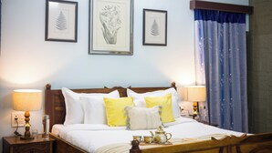A neatly made bed with white and yellow pillows, a bedside table with a lamp, and framed botanical prints on the wall. - Alaya Stays Abhimanyu Mansion Topaz 2BHk (Udaipur)