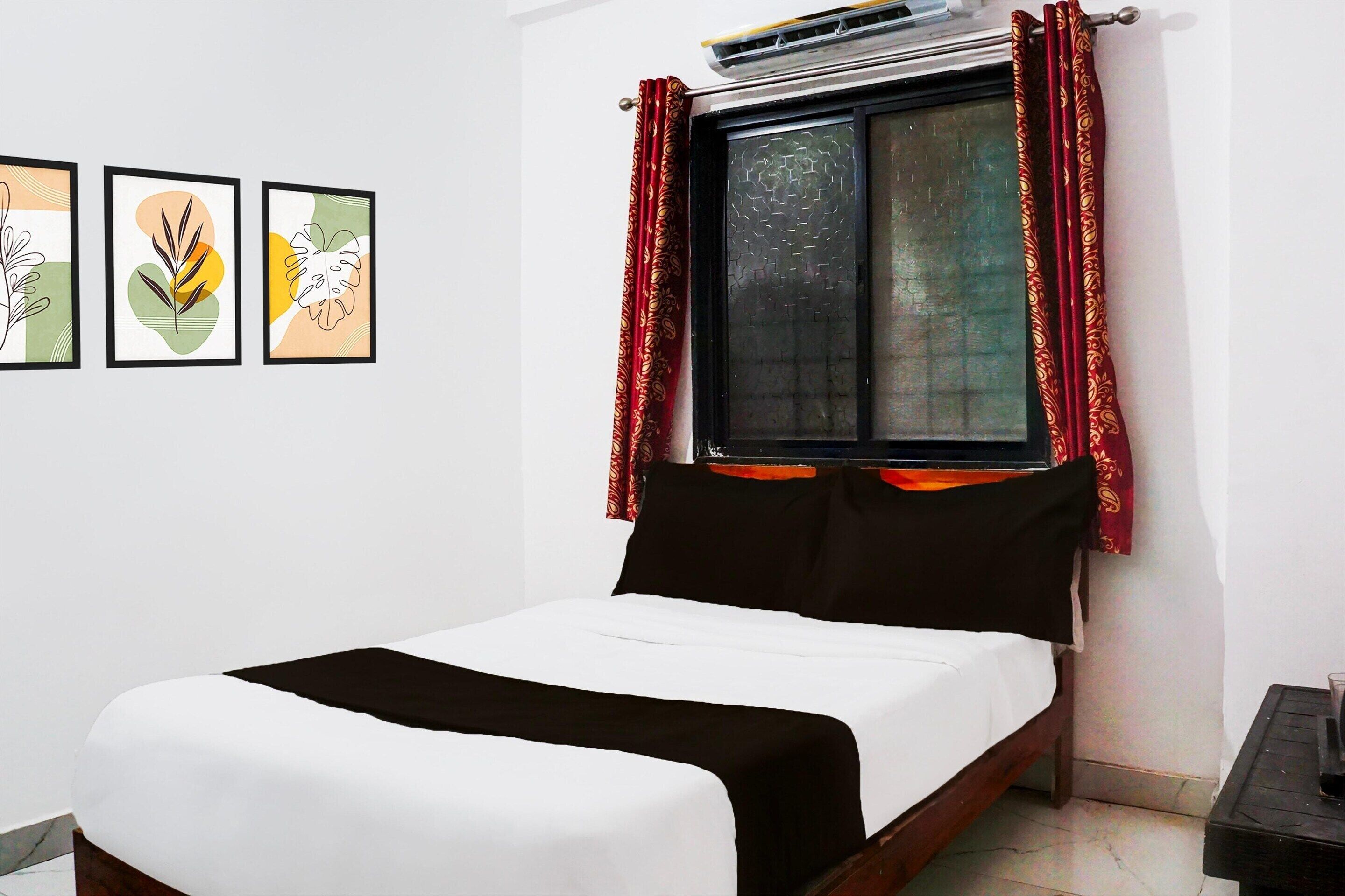 A bed with a white and black bedspread, a window with red curtains, and three framed artworks on the wall.