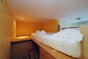 Ein kleines Zimmer mit einem Einzelbett, einem Holztisch und einer Bank, einem kleinen Regal und einem Deckenventilator.