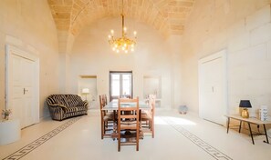 A spacious room with a high arched ceiling, a chandelier, a dining table with chairs, a sofa, and a small table with a lamp.