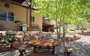 Eine Terrasse mit Holztisch und -stühlen, umgeben von Steinen und Pflanzen, mit einem Haus im Hintergrund.