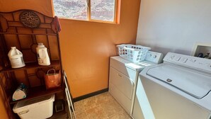 Un área de lavandería con lavadora, secadora, una ventana con vista a un paisaje rocoso y estantes con productos de limpieza.