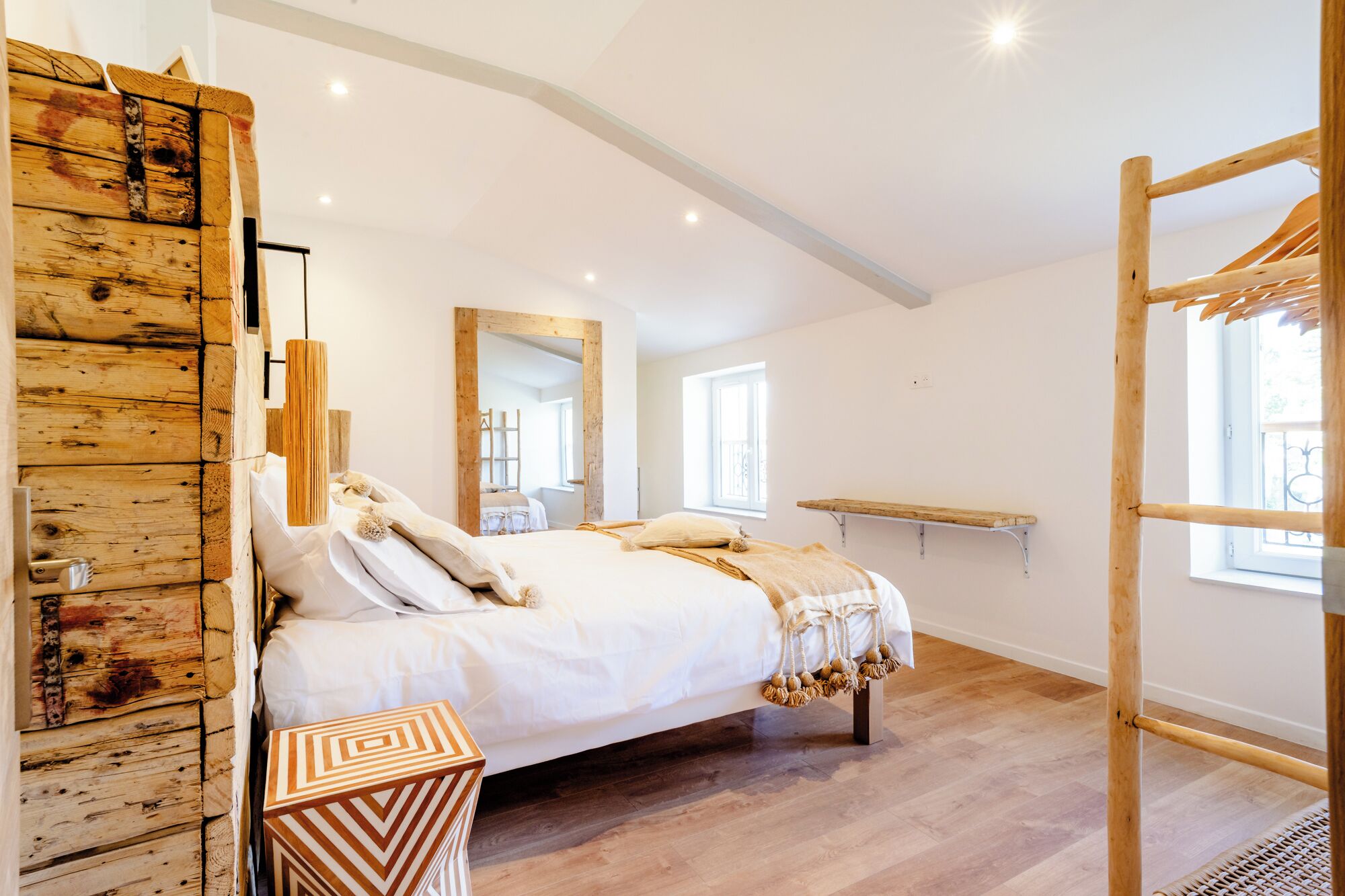 A bedroom with a wooden wall, a bed with white linens, a mirror, a window, and a wooden ladder.