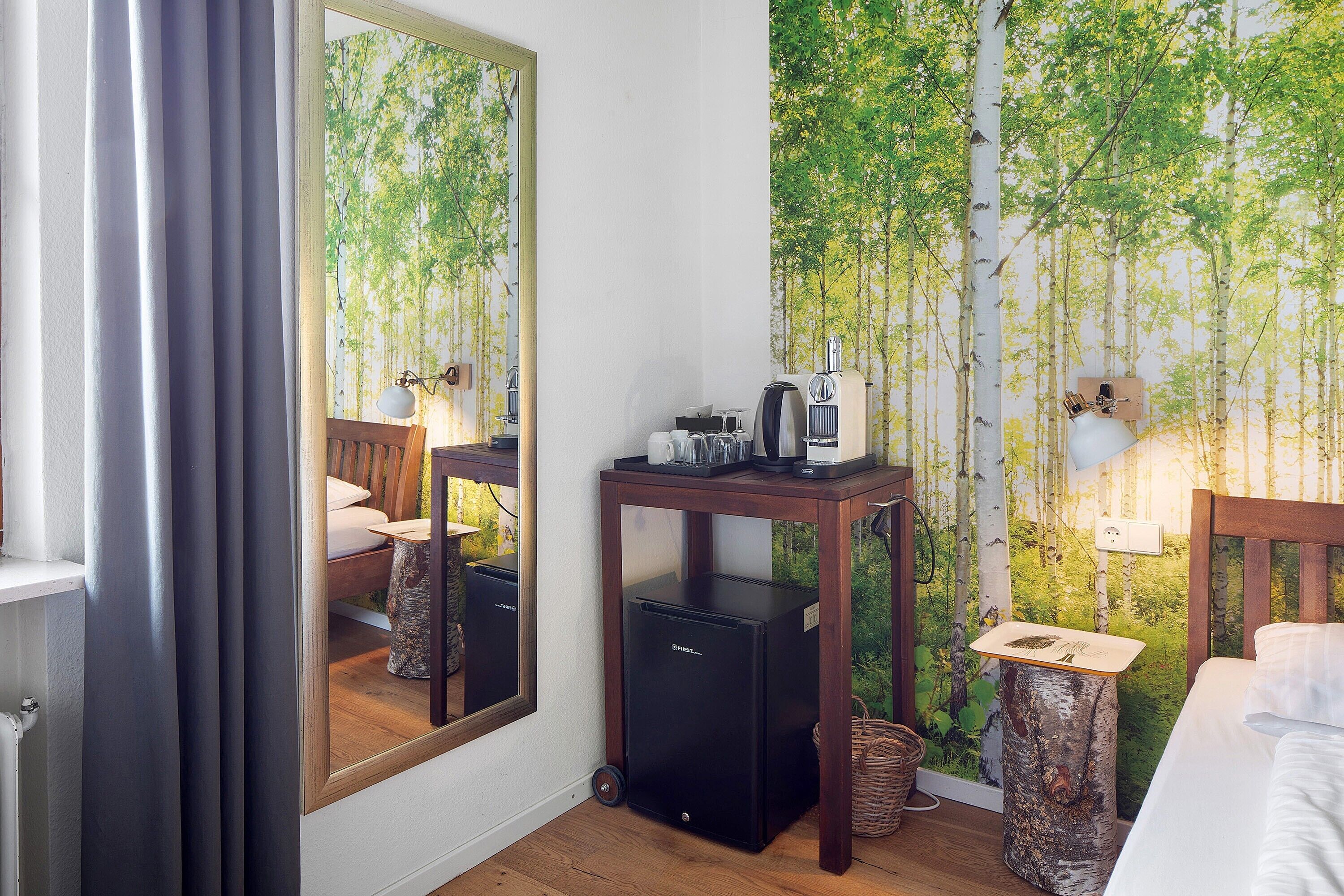 Une chambre d’hôtel avec un lit, un bureau doté d’une lampe, un petit réfrigérateur, un miroir et un mur recouvert d’un papier peint représentant un paysage naturel.