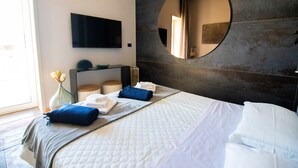 A modern hotel room with a large bed, a flat-screen TV mounted on the wall, a round mirror, and a vase with flowers. - C-Home Otranto – Design Apartment with Panoramic Terrace (Otranto)