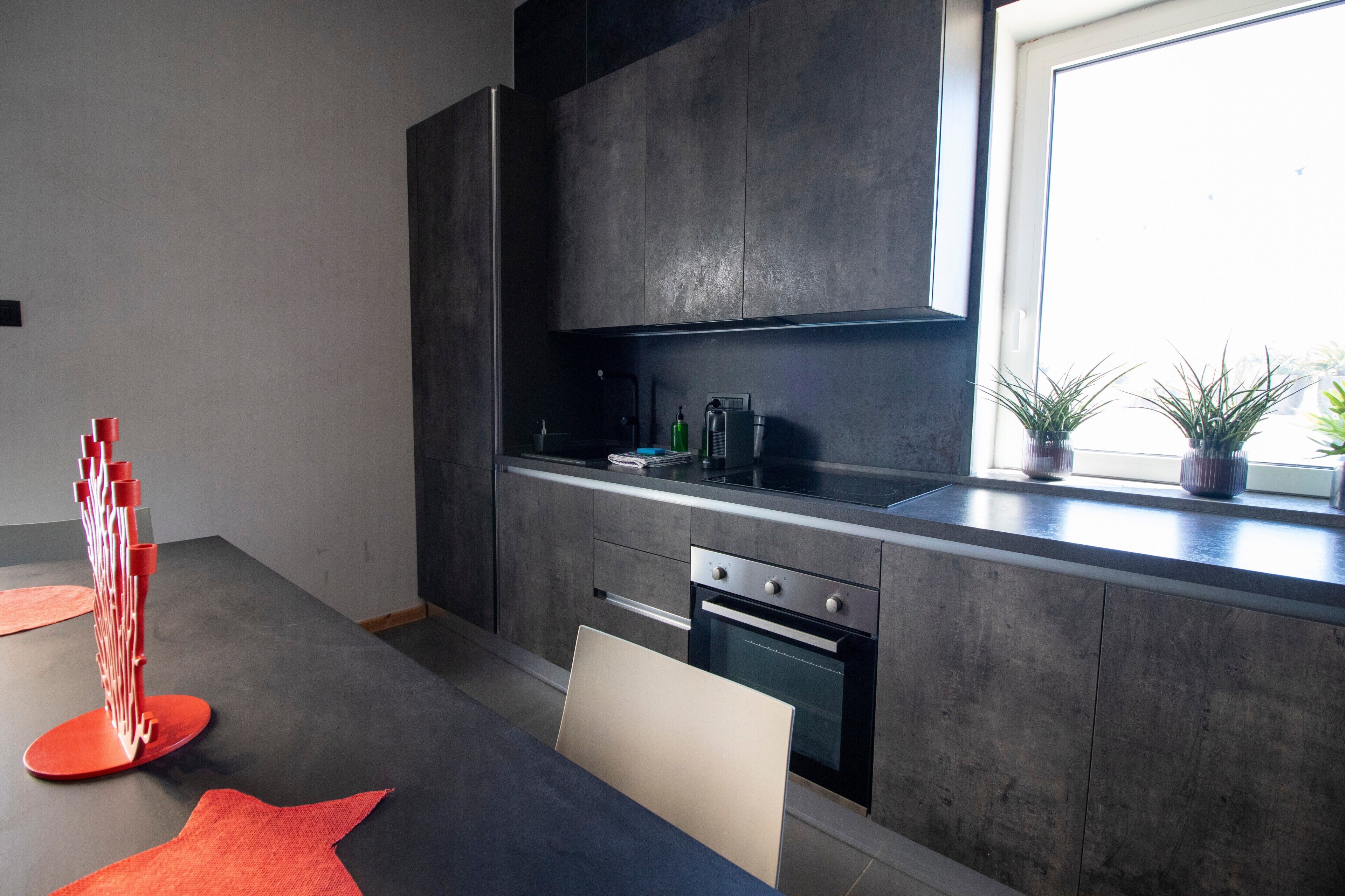 Eine moderne Küche mit dunklen Schränken, einem eingebauten Backofen und einem Fenster mit Pflanzen.