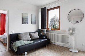 A modern living room with a black leather sofa, a standing fan, a window with curtains, and a radiator.