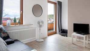 Uma sala de estar moderna com um sofá, um ventilador de chão, uma televisão sobre uma mesinha e uma vista através de uma janela grande.