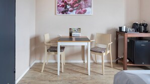 Ein Zimmer mit einem Holztisch und zwei Stühlen, einem kleinen Schreibtisch mit einem schwarzen Gerät sowie einem an der Wand hängenden Bild von rosafarbenen Blumen.