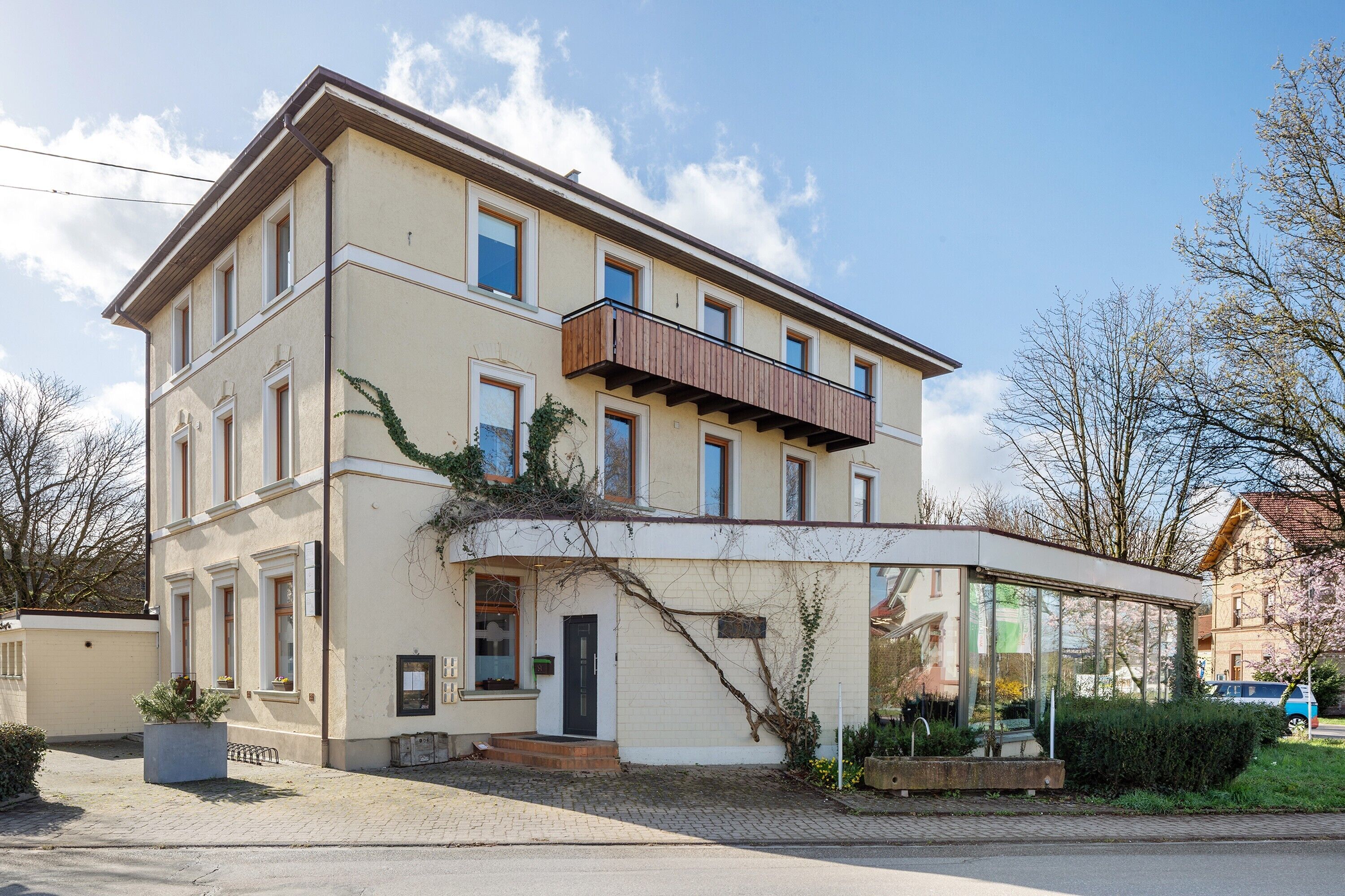 Un bâtiment à deux étages dot é d’un balcon, d’une véranda en verre et d’un espace d’entrée pavé.