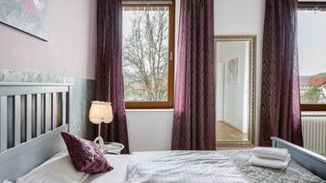 A bedroom with a bed, pillows, a bedside table, a lamp, and a window with curtains.