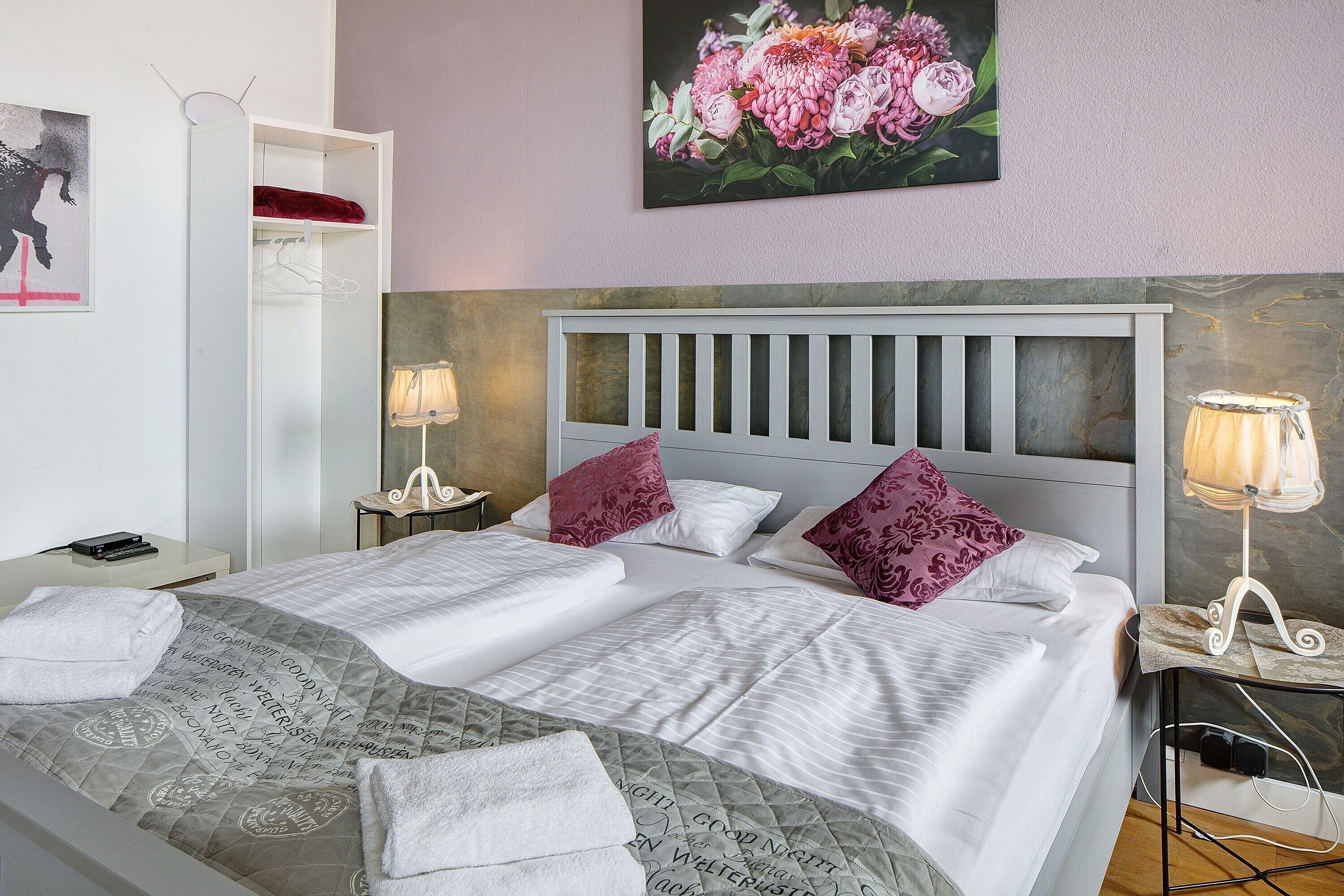 Een tweepersoonsbed met hoofdeinde, twee bedlampen, een nachtkastje, een garderobe en een bloemschilderij aan de muur.
