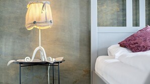 Una cama con ropa de cama blanca y una almohada morada, una mesita de noche con una lámpara, y una ventana con vistas.
