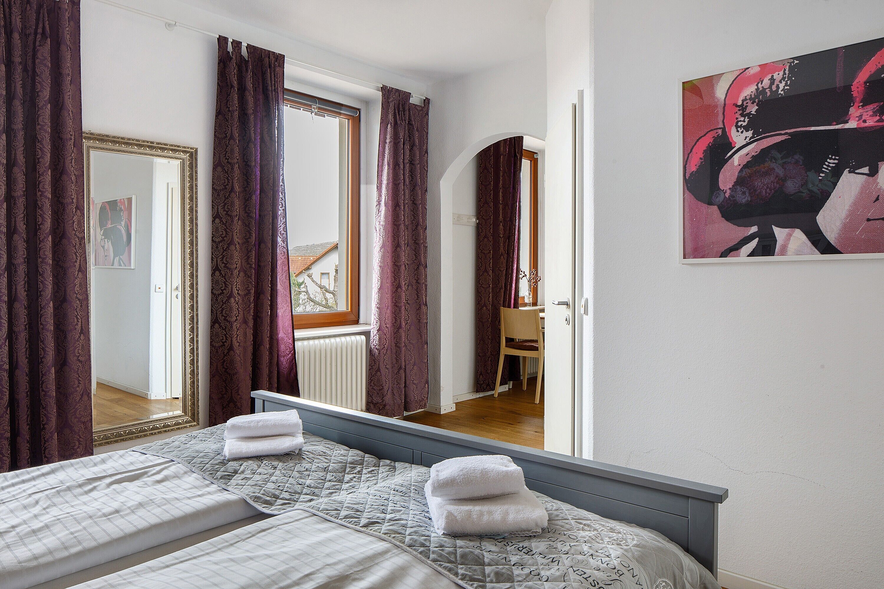 Una habitación de hotel con cama, espejo, ventana con cortinas, una silla y un cuadro en la pared.