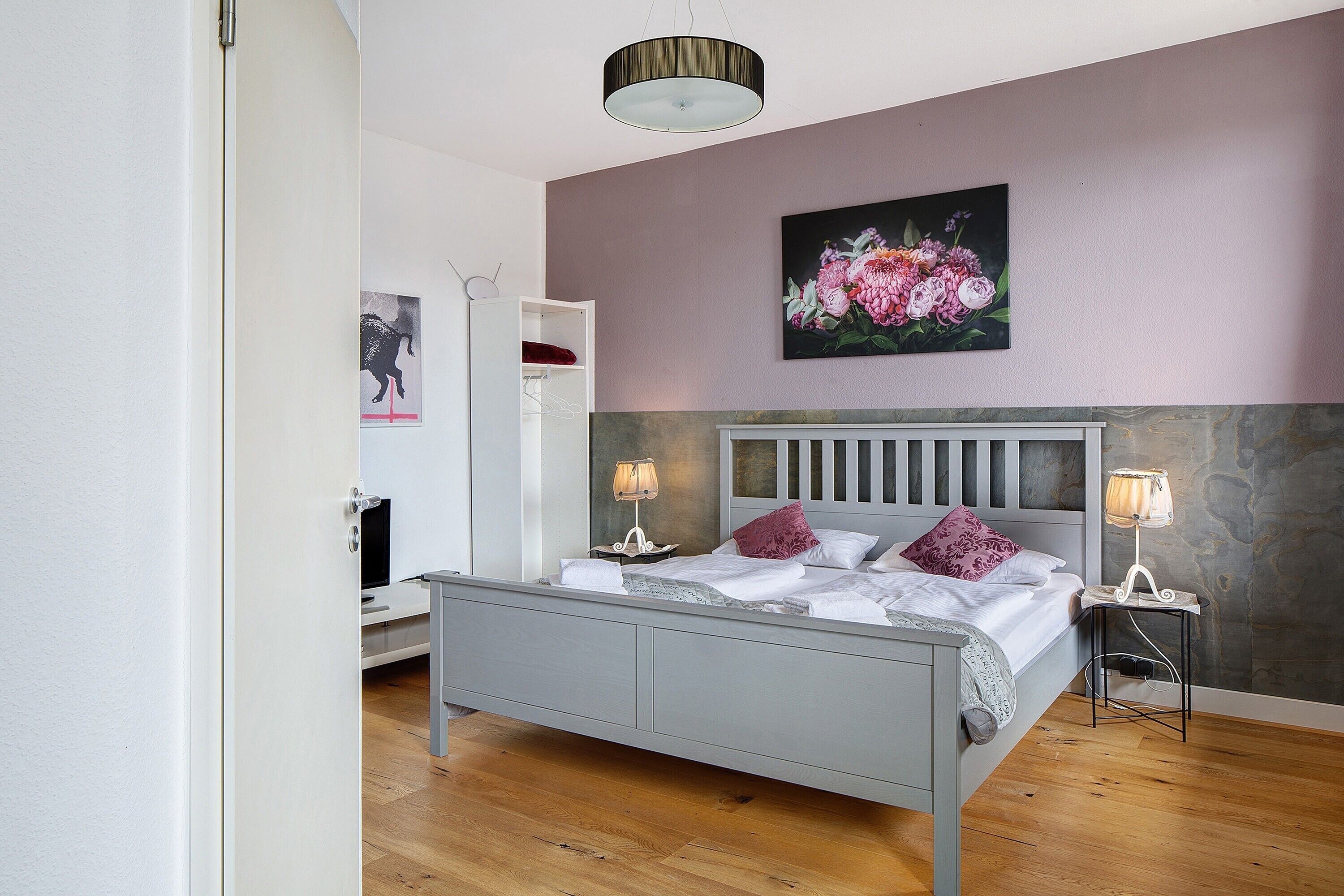 A bedroom with a bed, bedside lamps, a painting, and a wardrobe.