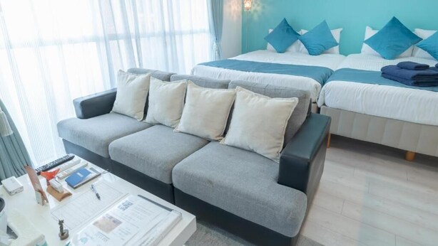 Habitación de hotel con sofá gris, cama con almohadas azules, mesita de noche con lámpara y ventana con cortinas translúcidas.