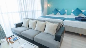 Habitación de hotel con sofá gris, cama con almohadas azules, mesita de noche con lámpara y ventana con cortinas translúcidas.
