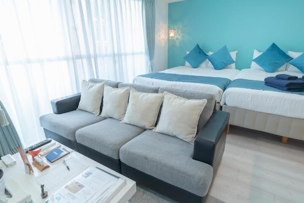 A hotel room with a grey sofa, a bed with blue pillows, a bedside table with a lamp, and a window with sheer curtains.