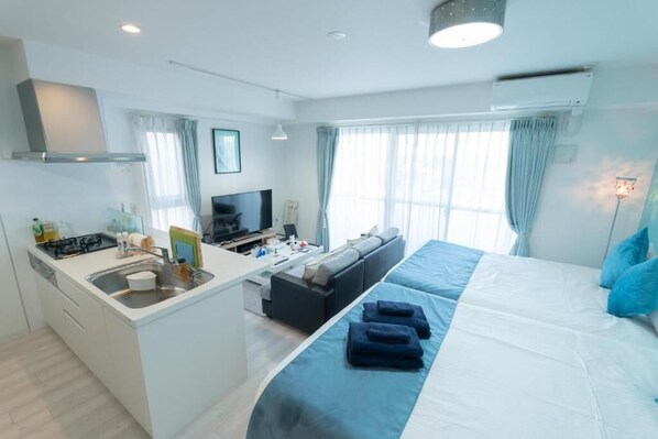 A modern hotel room with a kitchen area, a sofa, a bed with blue bedding, and a TV.