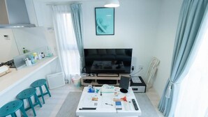 Una sala de estar moderna con un televisor de pantalla plana, una mesa de centro con varios objetos, una zona de cocina con estufa y un comedor con taburetes.