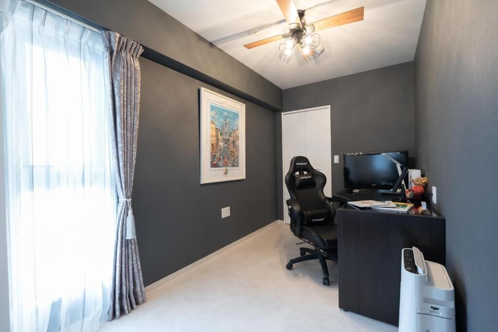 Ein Zimmer mit Deckenventilator, Schreibtisch mit Computer, Stuhl, Fernseher und einem gerahmten Bild an der Wand.