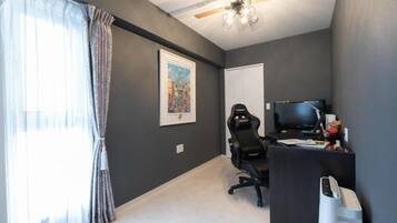 Ein Zimmer mit Deckenventilator, Schreibtisch mit Computer, Stuhl, Fernseher und einem gerahmten Bild an der Wand.