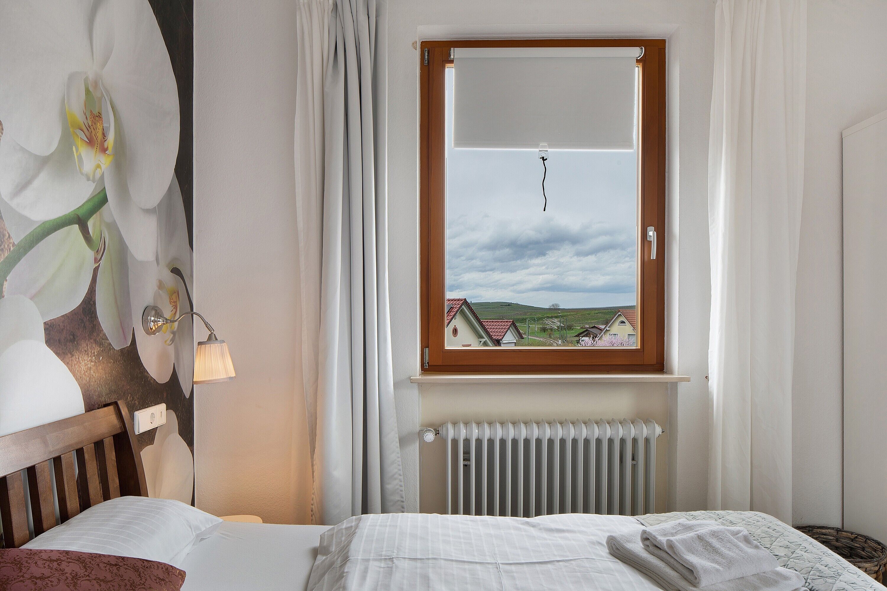 Ein Schlafzimmer mit einem Bett, einem Fenster mit Blick auf Häuser, einem Heizkörper und einer Wand mit einem Orchideenbild.