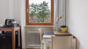 Een kamer met een houten tafel, een stoel, een kleine zwarte koelkast, een raam met uitzicht op bomen en een radiator.