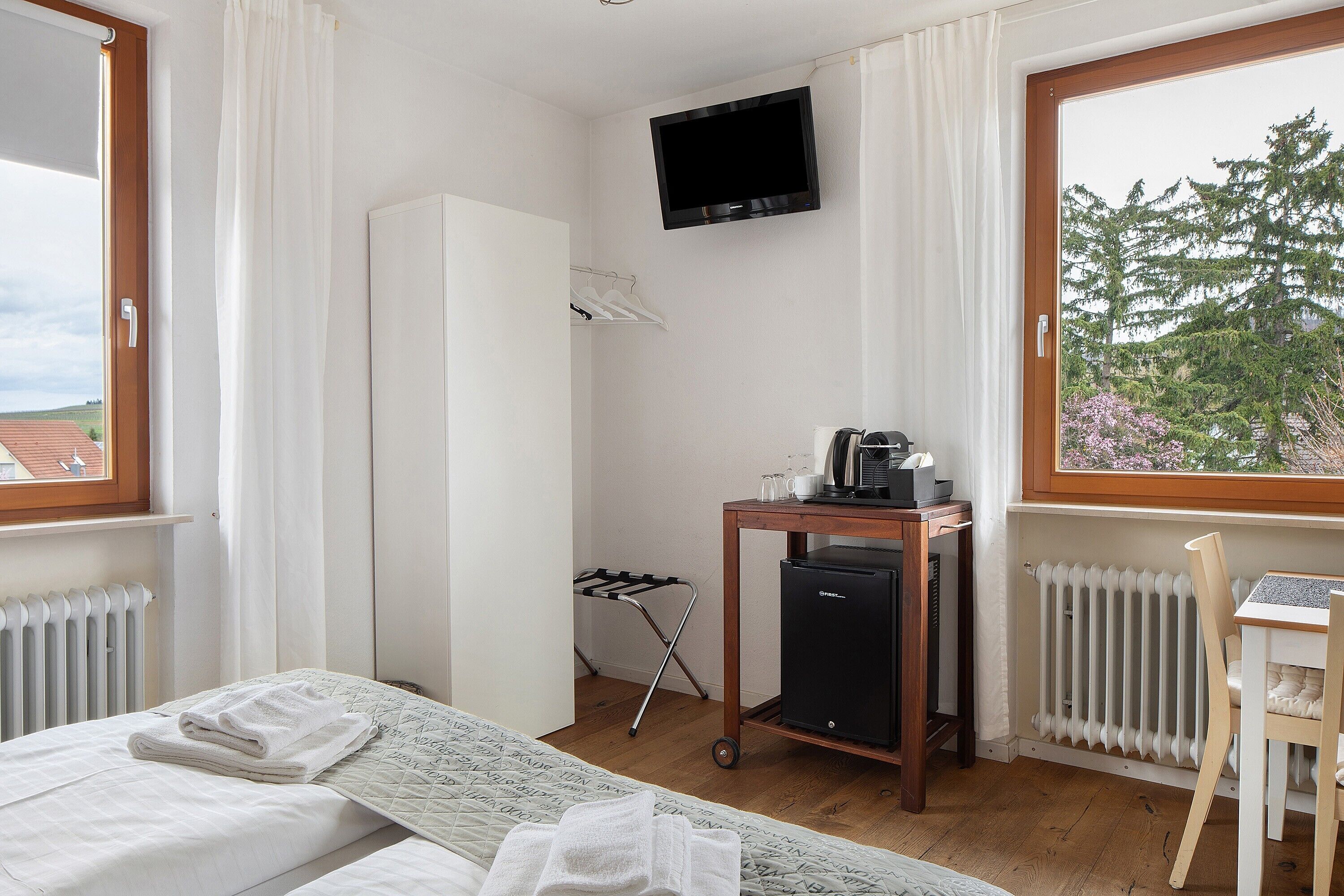 Ett hotellrum med en säng, ett skrivbord med en stol, ett element, en tv och ett fönster med utsikt över träd.