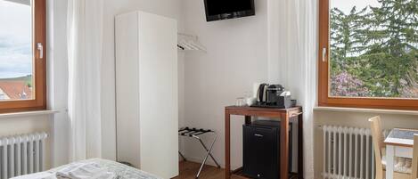 Ett hotellrum med en säng, ett skrivbord med en stol, ett element, en tv och ett fönster med utsikt över träd.