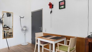 Una habitación con piso de madera, una mesa blanca con dos sillas, un ventilador de pie, una puerta y un espejo.