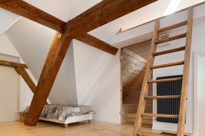 Une chambre à coucher aménagée dans un espace mansardé, avec des poutres apparentes, un lit en hauteur, une échelle en bois et un radiateur.