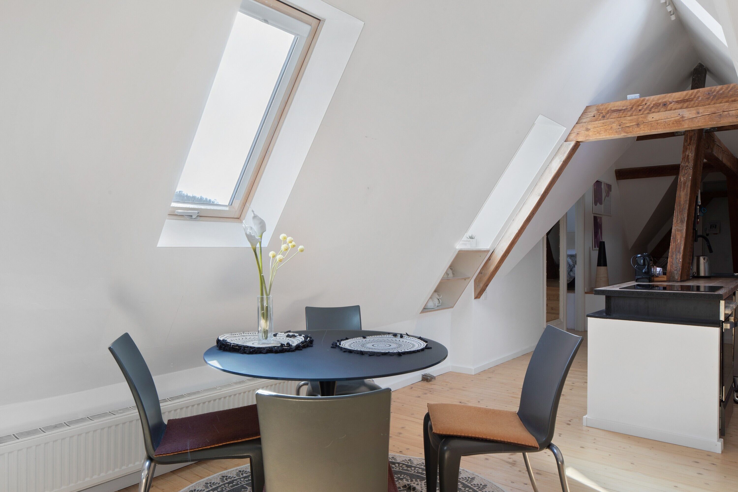 A dining area with a round table, chairs, a vase with flowers, and a view of the sky through a skylight.