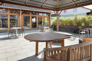 Une terrasse extérieure avec des tables et des chaises en bois, un espace salon couvert et de grandes fenêtres.